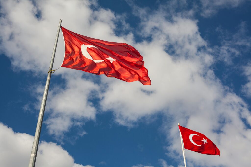 Two Turkish flags waving in the wind against a bright blue sky filled with white clouds.